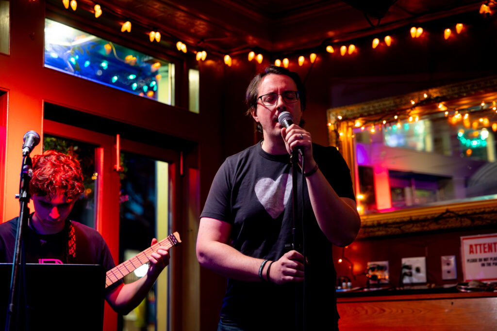 A singer holding a microphone performs on a small, warmly lit stage while a guitarist plays beside him, surrounded by string lights and colorful reflections.