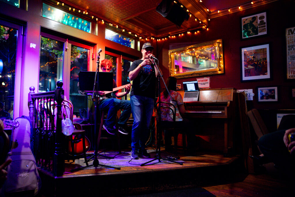 Photo of a small live music performance in an intimate venue, with a singer at a microphone, a guitarist seated beside him, and a pianist at an upright piano under string lights.