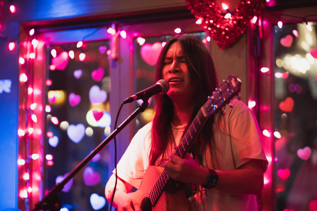 Singer-songwriter performing live indoors, singing into a microphone while playing an acoustic guitar, surrounded by pink heart-shaped lights and warm stage lighting.
