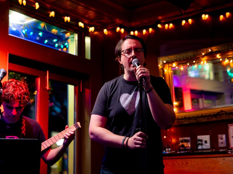 A singer holding a microphone performs on a small, warmly lit stage while a guitarist plays beside him, surrounded by string lights and colorful reflections.