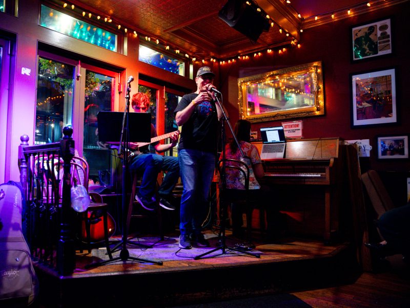 Photo of a small live music performance in an intimate venue, with a singer at a microphone, a guitarist seated beside him, and a pianist at an upright piano under string lights.